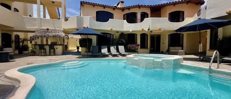 Una piscina con sillones de baño y sombrillas, rodeada de edificios con balcones.
