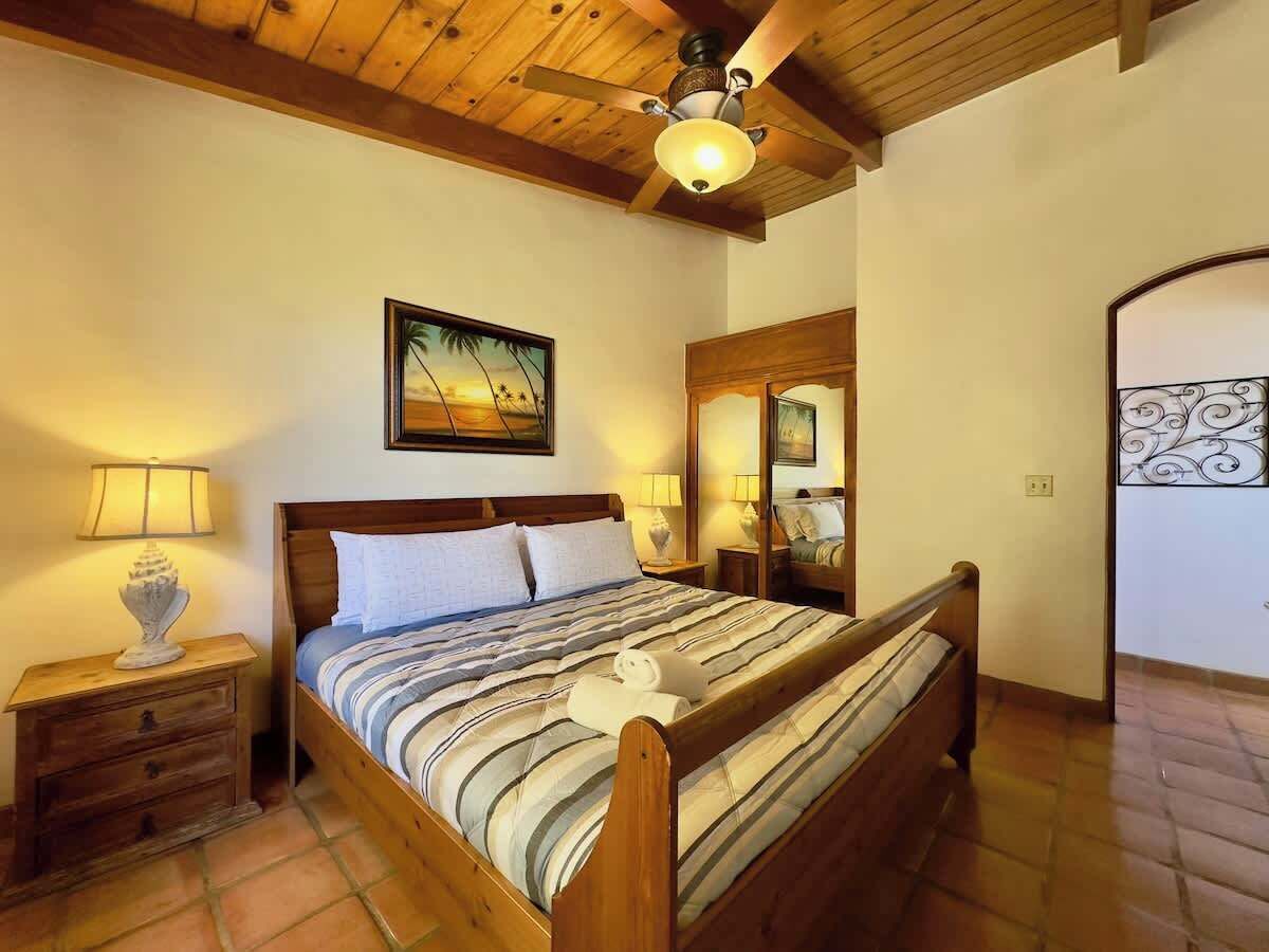 A wooden bed with a striped bedspread, a nightstand with a lamp, a ceiling fan, and a painting on the wall.