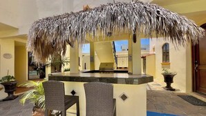 An outdoor bar area with a thatched roof, two wicker chairs, and potted plants.