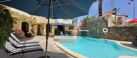 A swimming pool with lounge chairs and umbrellas, surrounded by stone walls and palm trees.