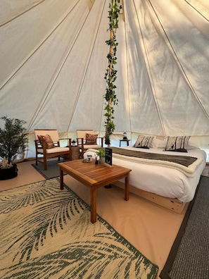 A tent interior with a bed, wooden furniture, a hanging plant, and a welcome sign.