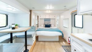 A compact RV interior with a bed, seating area, kitchenette, and a small table.