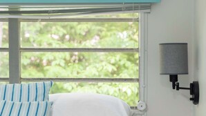 A bedroom with a bed, a window with blinds, a nightstand with a lamp, and a view of greenery outside.