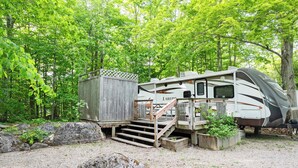 A recreational vehicle (RV) parked in a wooded area with a wooden deck and steps leading to a covered structure.