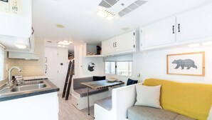 A compact kitchenette with a sink, stove, and cabinets, a small dining table, and a sofa with cushions.
