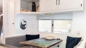 A compact RV interior with a kitchenette, a dining area with a wooden table, and a window with a lattice screen.