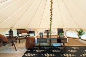 Interior de una carpa con una cama, una mesa con una planta, una silla, una mesita con un farol y un letrero de bienvenida.