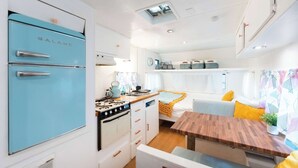 A compact RV kitchen with a blue refrigerator, stove, and sink, a wooden dining table, and a cozy seating area with cushions. - Blue Glamping RV w/ Nordic Spa Access near Grotto (Miller Lake)