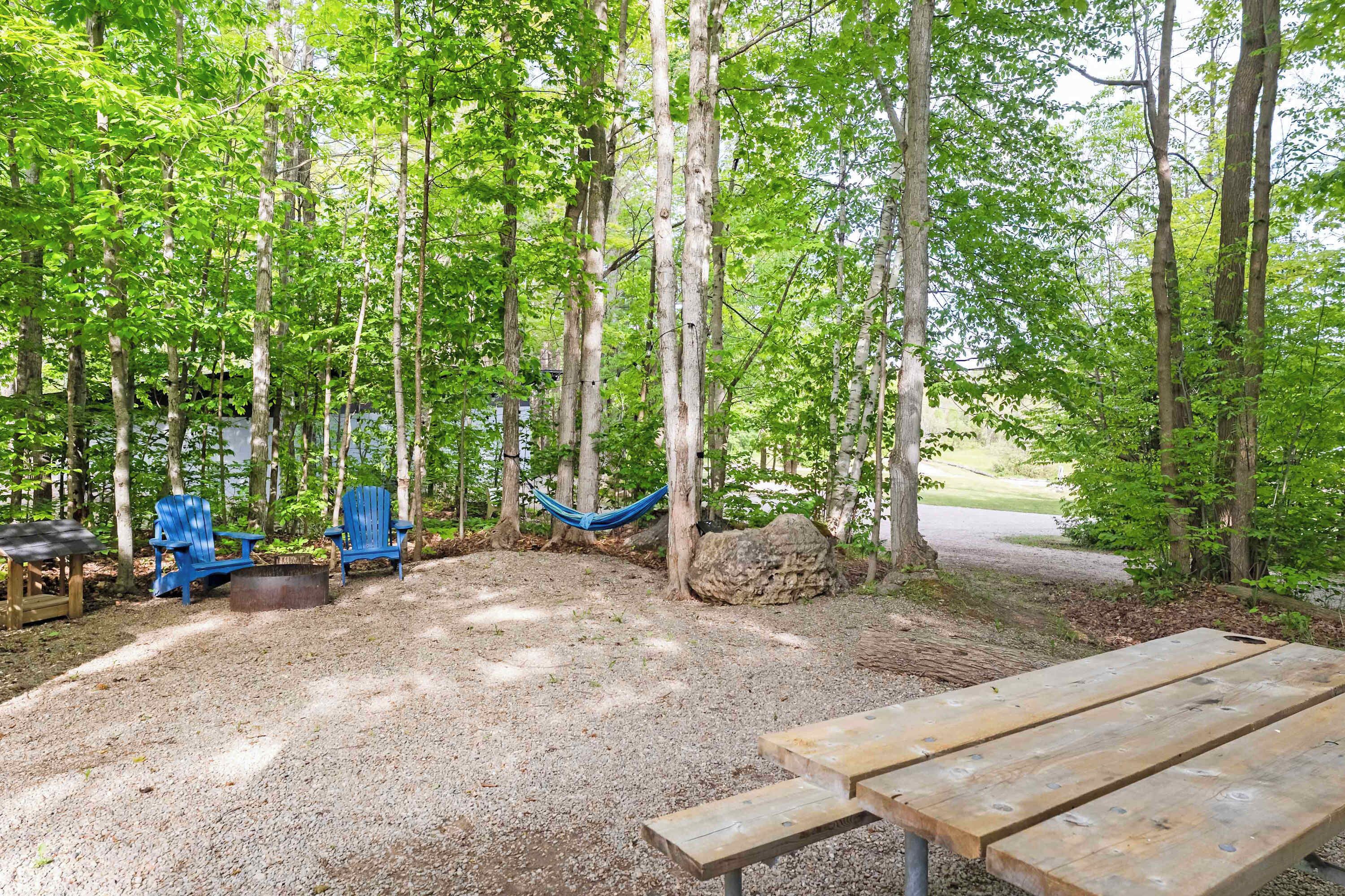 A forested campsite with a picnic table, blue Adirondack chairs, and a hammock.