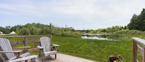Een houten terras met Adirondack-stoelen met uitzicht op een rustig meer, omgeven door groen.