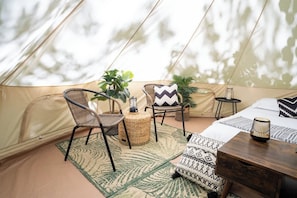 A tent interior with a bed, a dining table, chairs, a small table with a lamp, and potted plants.