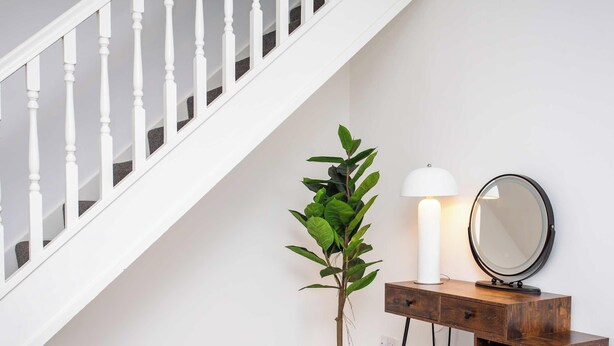 Eine Treppe mit weißen Balustraden, ein Blumentopf, eine weiße Tischlampe und ein runder Spiegel auf einer Holzkonsole.