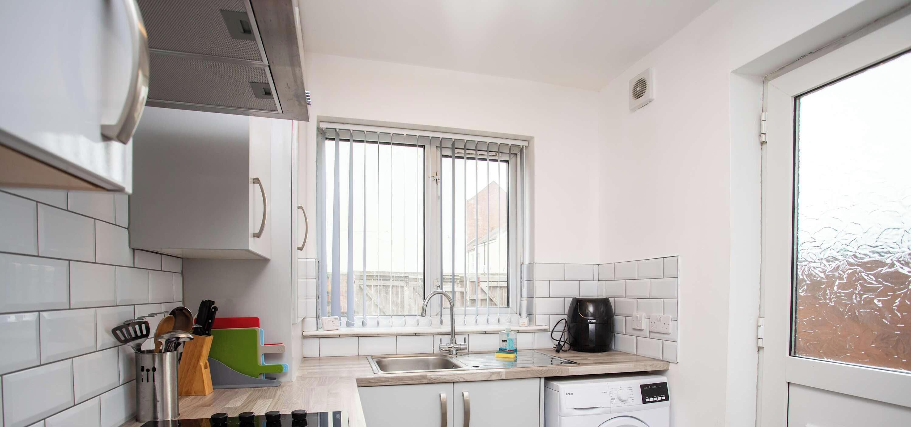 Une cuisine compacte avec des carreaux blancs, une fenêtre avec des stores, un évier, une machine à café et un lave-linge.