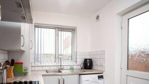 Eine kleine Küche mit weißen Fliesen, einem Fenster mit Jalousien, einem Spülbecken, einer Kaffeemaschine und einer Waschmaschine.