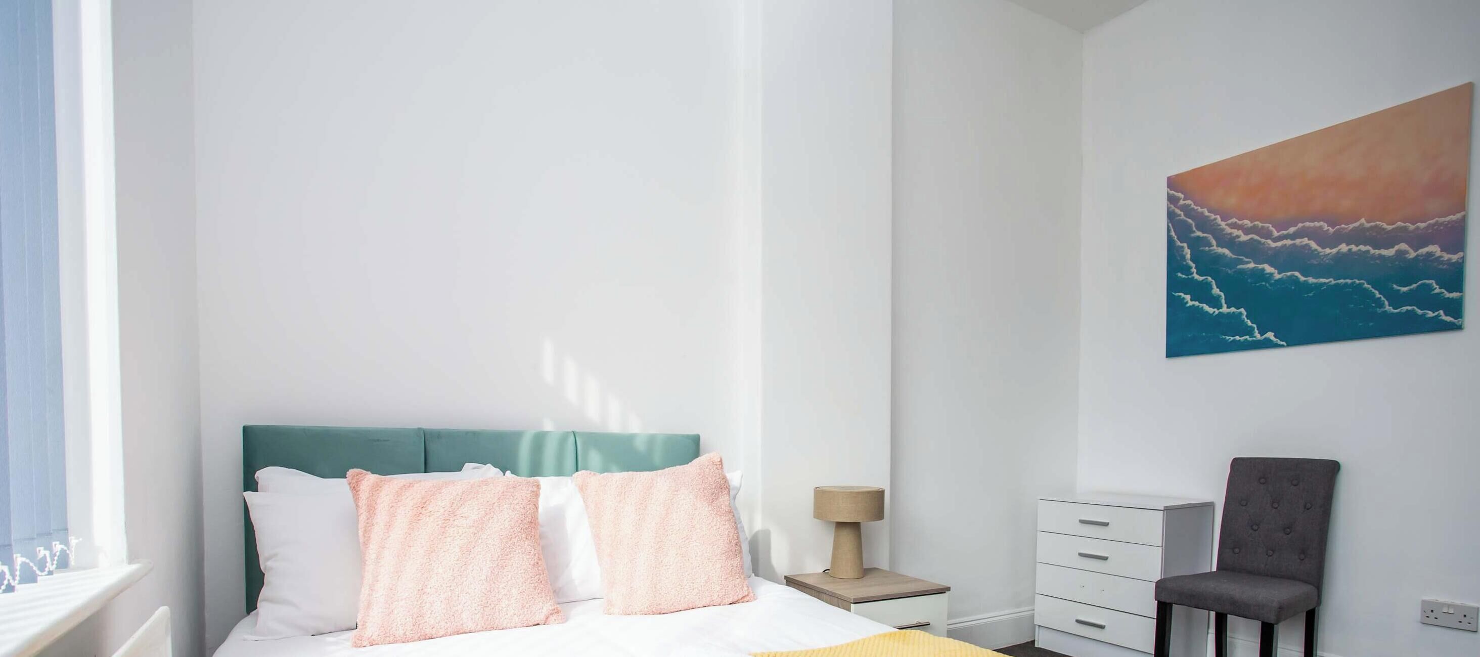 Ein Schlafzimmer mit Bett, Kissen, Nachttisch, Stuhl und einem Bild an der Wand.