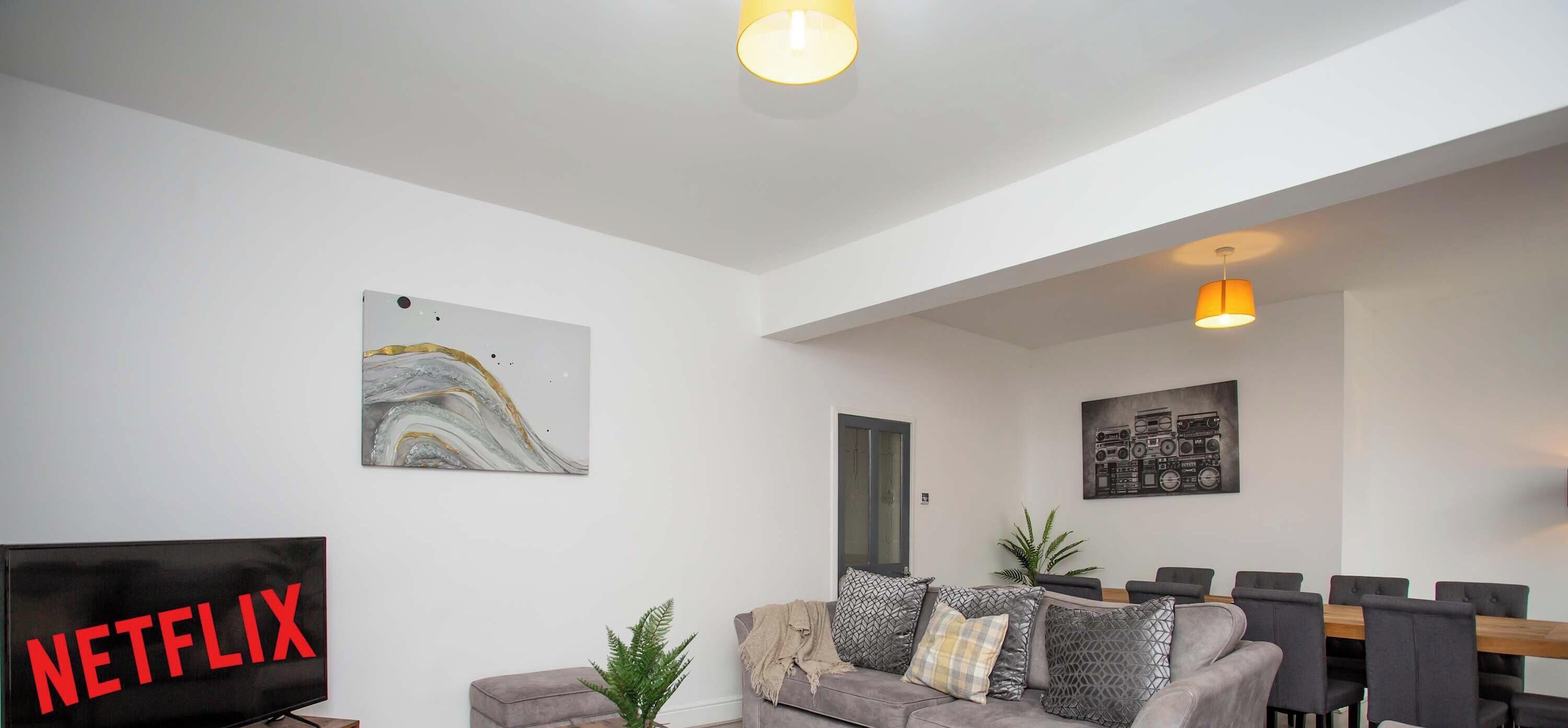 A modern living room with a flat-screen TV, a gray sofa with patterned cushions, a dining table with chairs, and two wall-mounted pendant lights.