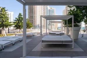 An outdoor seating area with white tables and benches, surrounded by tall buildings and trees.