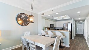 A modern kitchen with white cabinetry, a central island, and a dining area with a wooden table and white chairs. - Seawind 1502 | Beach Front (Gulf Shores)