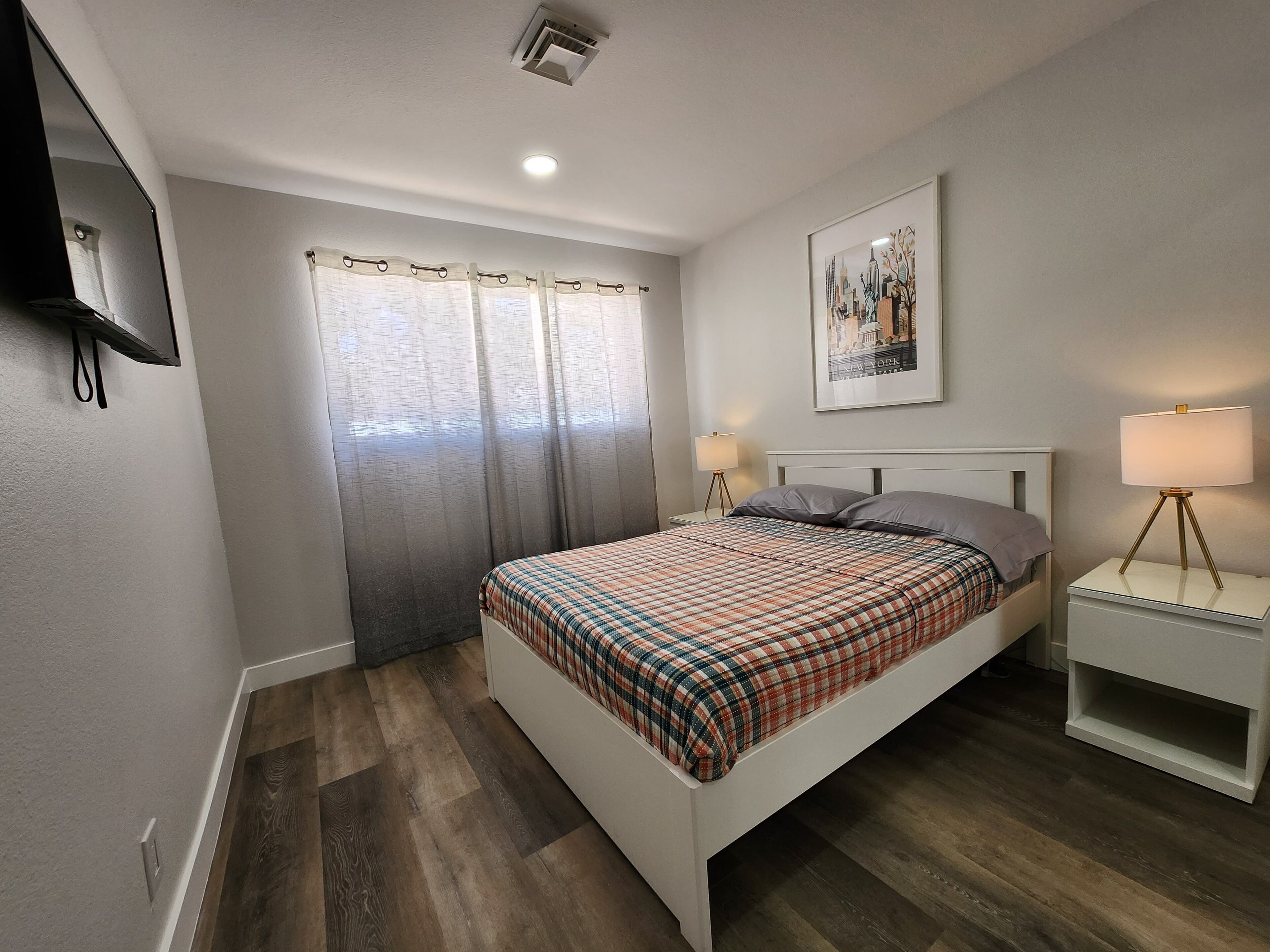 A modern bedroom with a bed, bedside lamps, a mounted TV, and a framed cityscape picture.