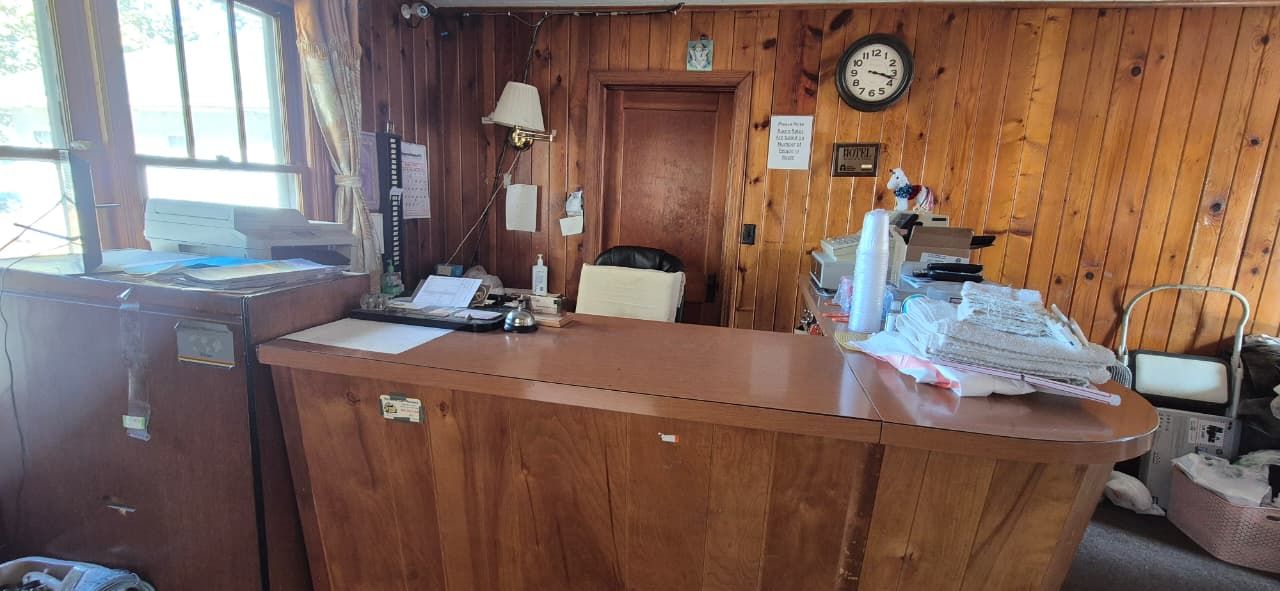 A wooden reception desk with a computer, papers, and a lamp. There is a chair and a clock on the wall.