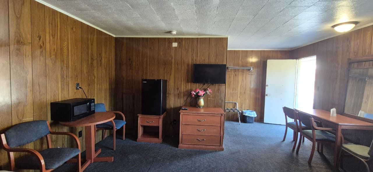 Quarto com paredes em painel de madeira, televisão de tela plana na parede, cômoda de madeira com gavetas, mesa pequena com cadeira e mesa de jantar com cadeiras.