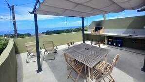 An outdoor patio with a table and chairs, a grill, and a view of the ocean. - Charming Rustic House in Nordeste surrounded by nature (Nordeste)