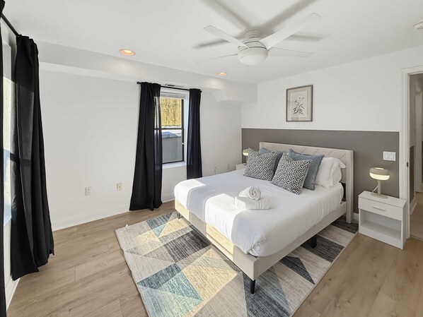 Un dormitorio con cama, mesita de noche, ventilador de techo y ventana con cortinas.