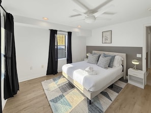Un dormitorio con cama, mesita de noche, ventilador de techo y ventana con cortinas.