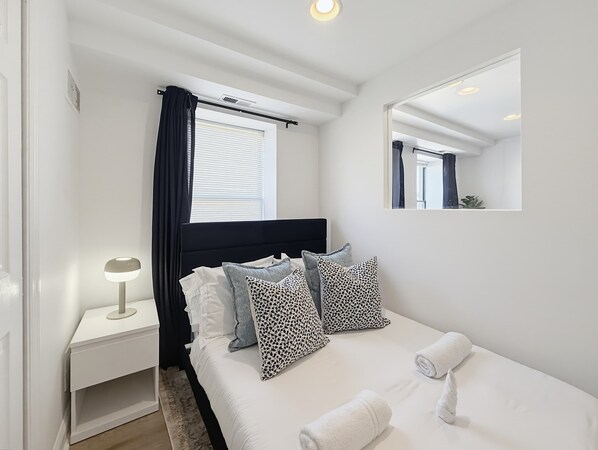 Habitación de hotel con cama, mesita de noche con lámpara, ventana con cortinas, espejo y luz en el techo.