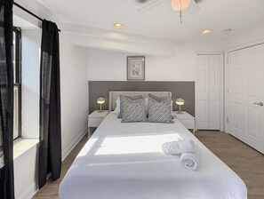 Habitación de hotel con una cama grande, dos mesitas de noche con lámparas, un cuadro enmarcado en la pared y un ventanal con cortinas.