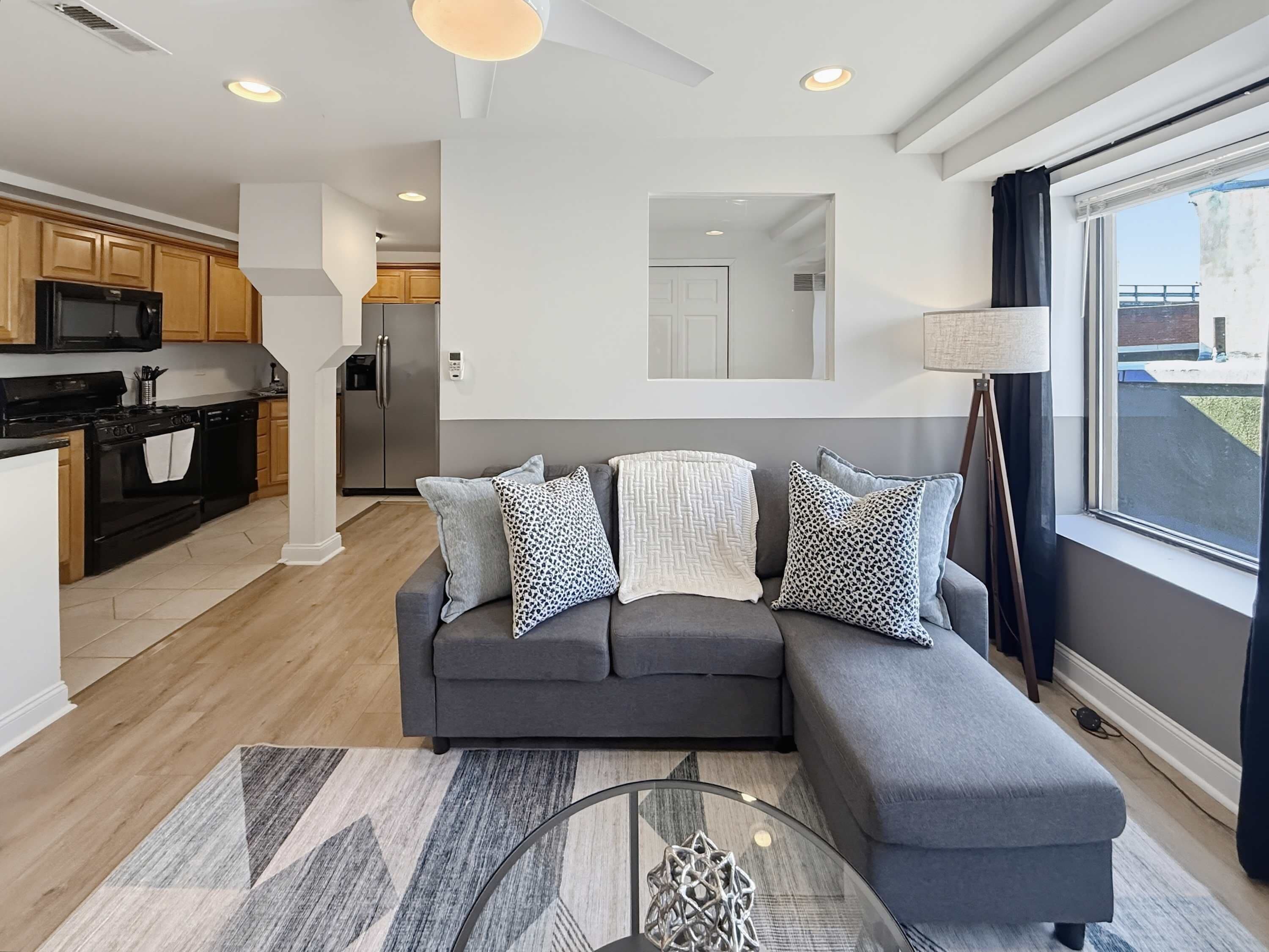 A modern living room with a grey sofa, patterned cushions, a glass coffee table, and a view of the kitchen and balcony.