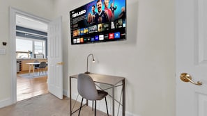 A modern room with a flat-screen TV displaying a streaming service interface, a desk with a chair, and a door leading to a kitchen area. - Charming 2BD by Independence Hall | Sleeps 6 (Philadelphia)