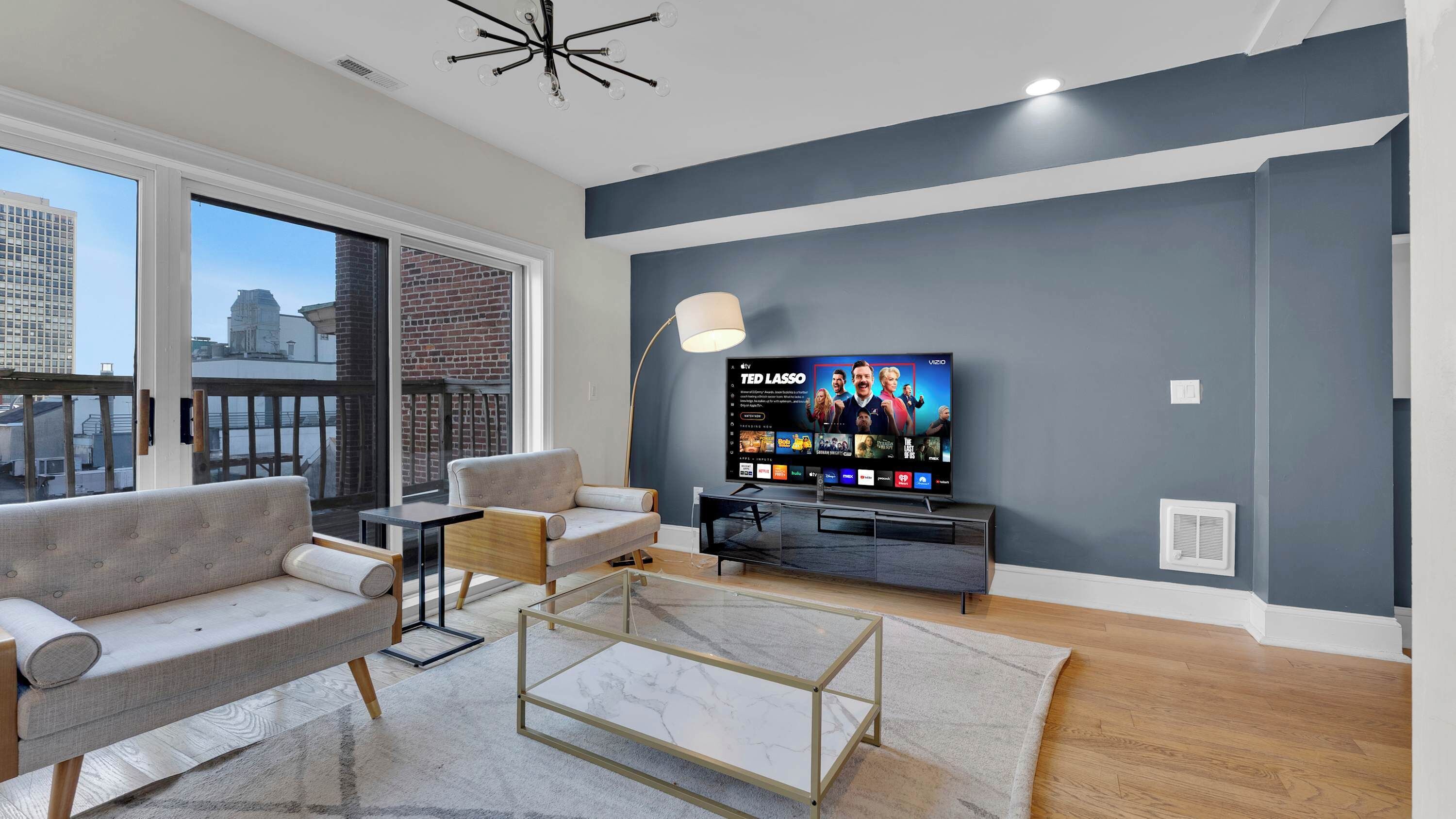 A modern living room with a large flat-screen TV, comfortable seating, a glass coffee table, and a view of the city through large windows.