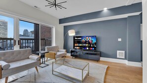 A modern living room with a large flat-screen TV, comfortable seating, a glass coffee table, and a view of the city through large windows.