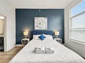 A bedroom with a bed, two bedside tables, a wall-mounted lamp, a large floral artwork, and a window with blinds.