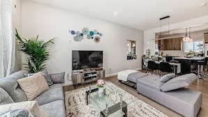 A modern living room with a sofa, a coffee table, and a TV. There is a kitchen area with wooden cabinets and a dining table. - Cozy beautiful house (Calgary)
