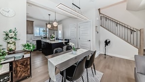 A modern kitchen with a dining area, featuring a long table, chairs, pendant lights, and a staircase. - Cozy beautiful house (Calgary)