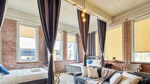 Une chambre d’hôtel moderne avec un mur en briques, de grandes fenêtres, un canapé, un lit et une petite table.