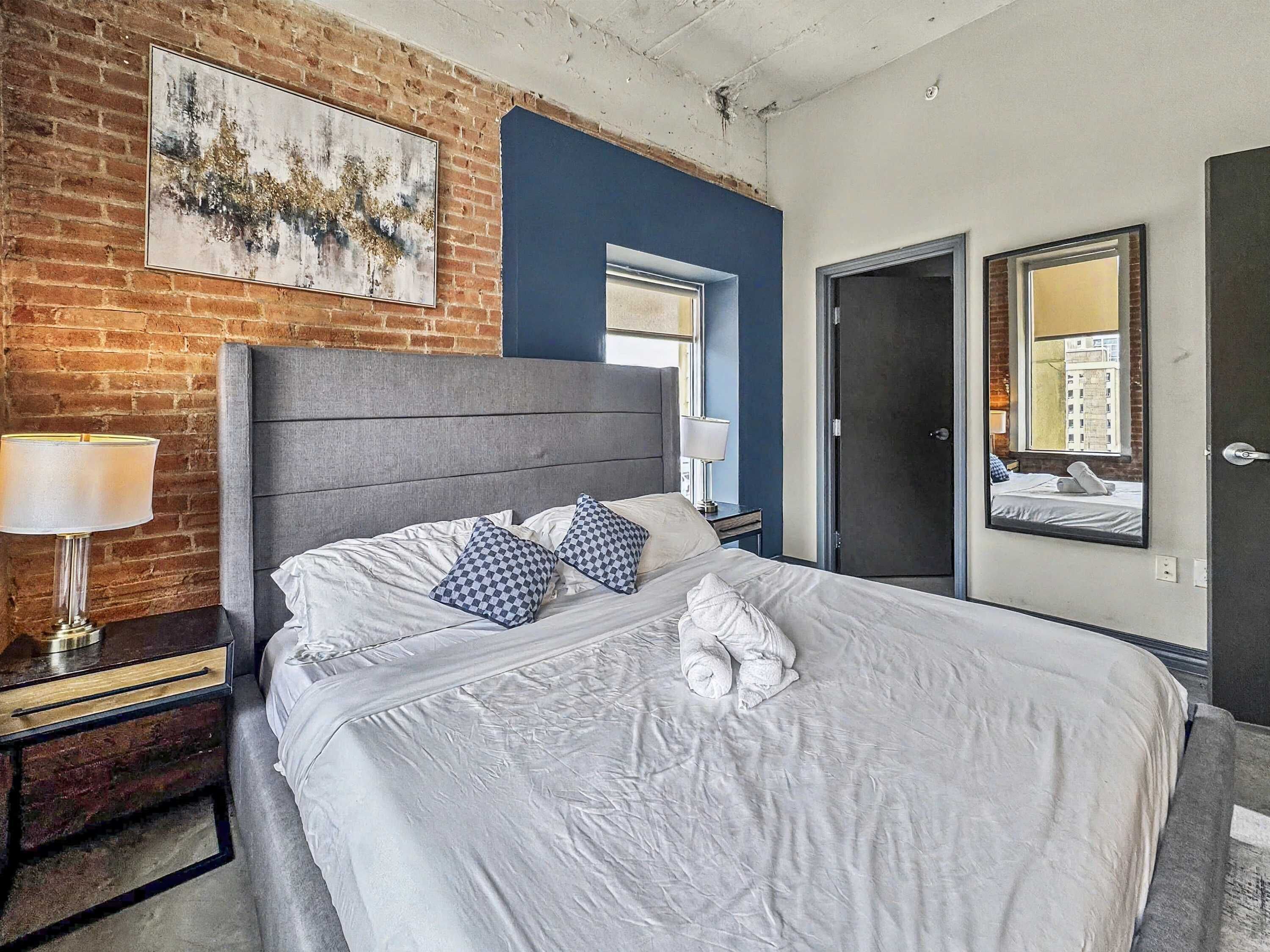 A hotel room with a brick wall, a large bed with white bedding, checkered pillows, a nightstand with a lamp, and a window with a view of buildings.