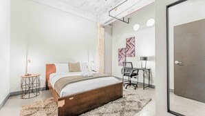 Une chambre moderne avec un lit en bois, une table de chevet ronde avec une lampe, un bureau avec une chaise et des œuvres d’art abstraites accrochées au mur.