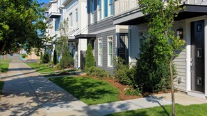 A row of townhouses with a sidewalk, small trees, and a clear sky.
