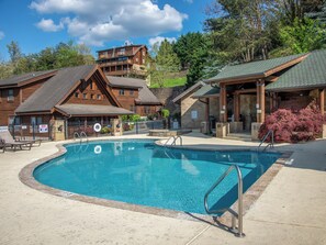 A pool area with a wooden lodge in the background, a covered seating area, and a sign indicating shallow water. - Up To Par 3211 by Jackson Mountain Rentals (Pigeon Forge)