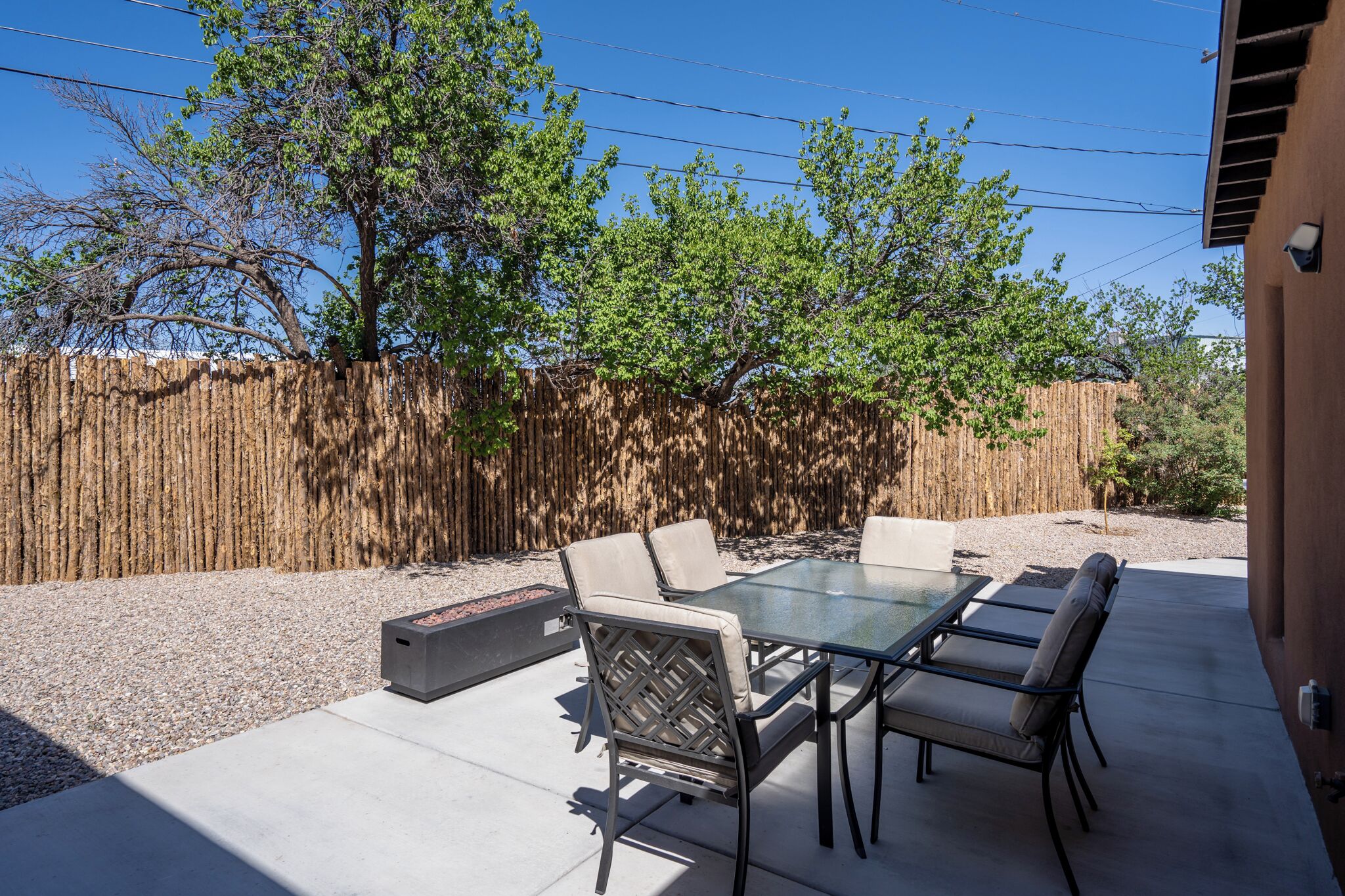 A patio with a glass table and chairs, surrounded by a wooden fence and trees.