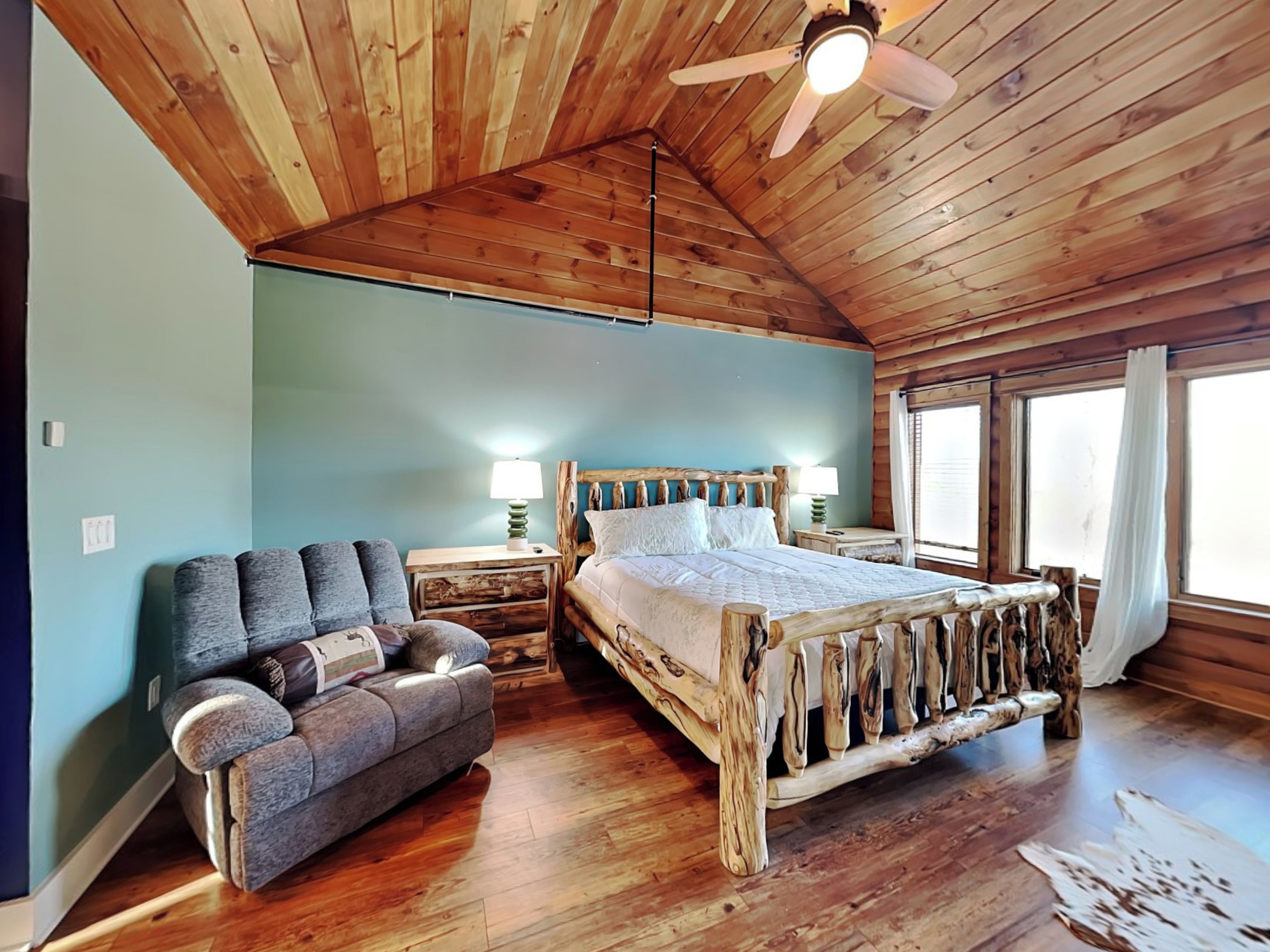 A rustic cabin bedroom with a wooden ceiling, a bed with a unique wooden headboard, a gray sofa, and a nightstand with lamps.