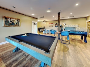 A pool table in a room with a wooden floor, a foosball table, and a bar area.