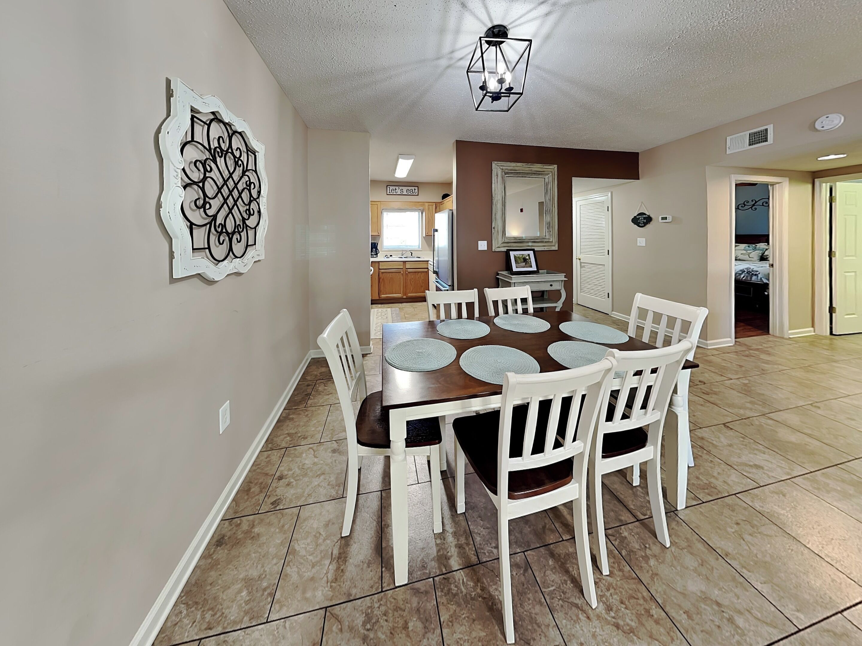 A dining area with a wooden table and white chairs, a kitchen in the background, and a decorative wall piece.