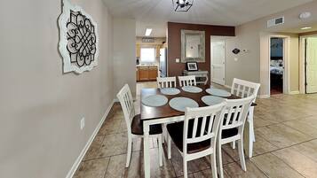 A dining area with a wooden table and white chairs, a kitchen in the background, and a decorative wall piece.