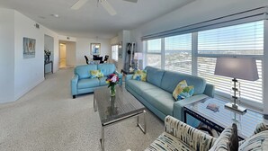 A spacious living room with a large window, a ceiling fan, and a dining area in the background.