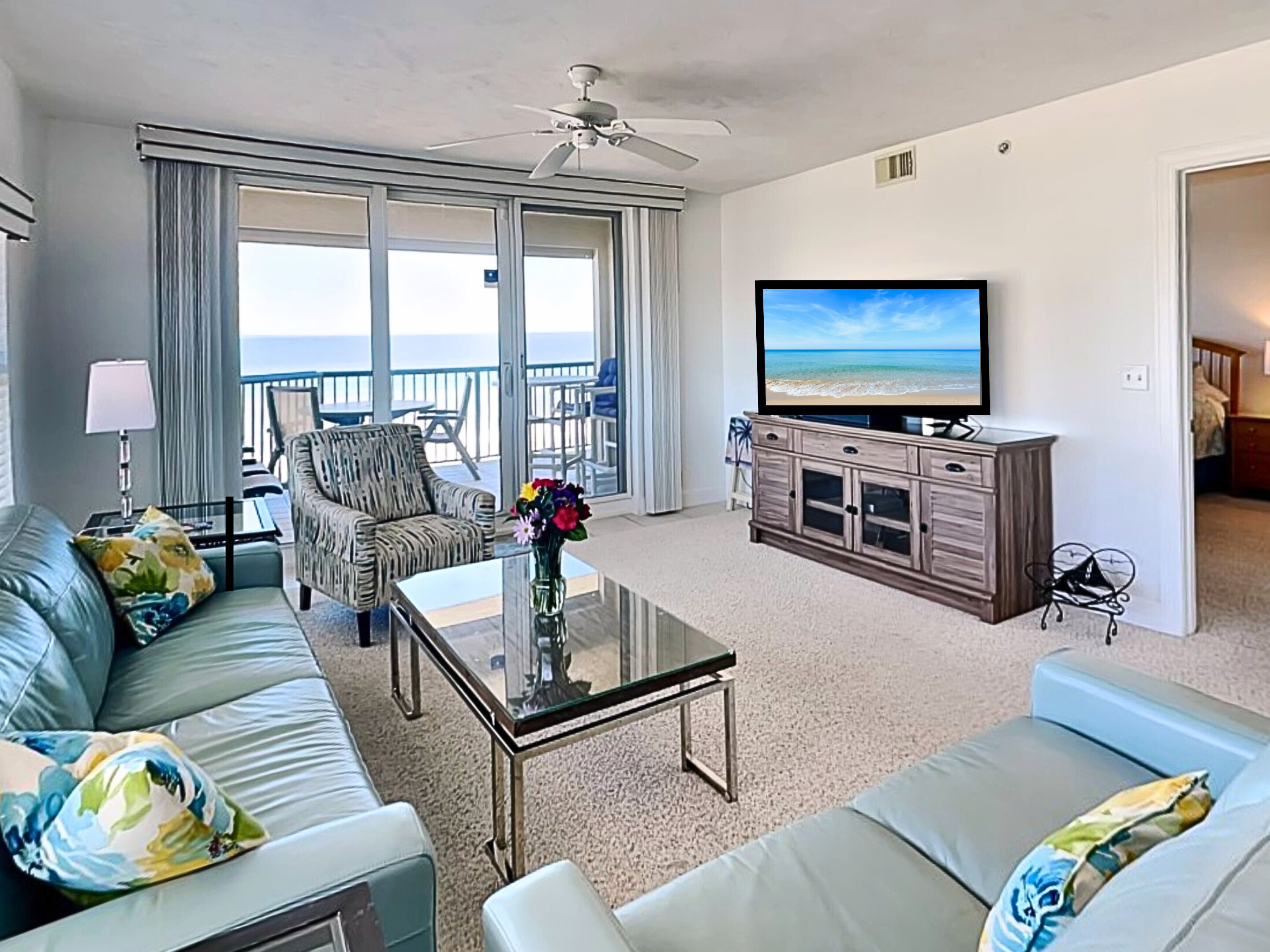 A living room with a sofa, armchair, and a coffee table. There is a television on a cabinet, and a balcony with a view of the ocean.