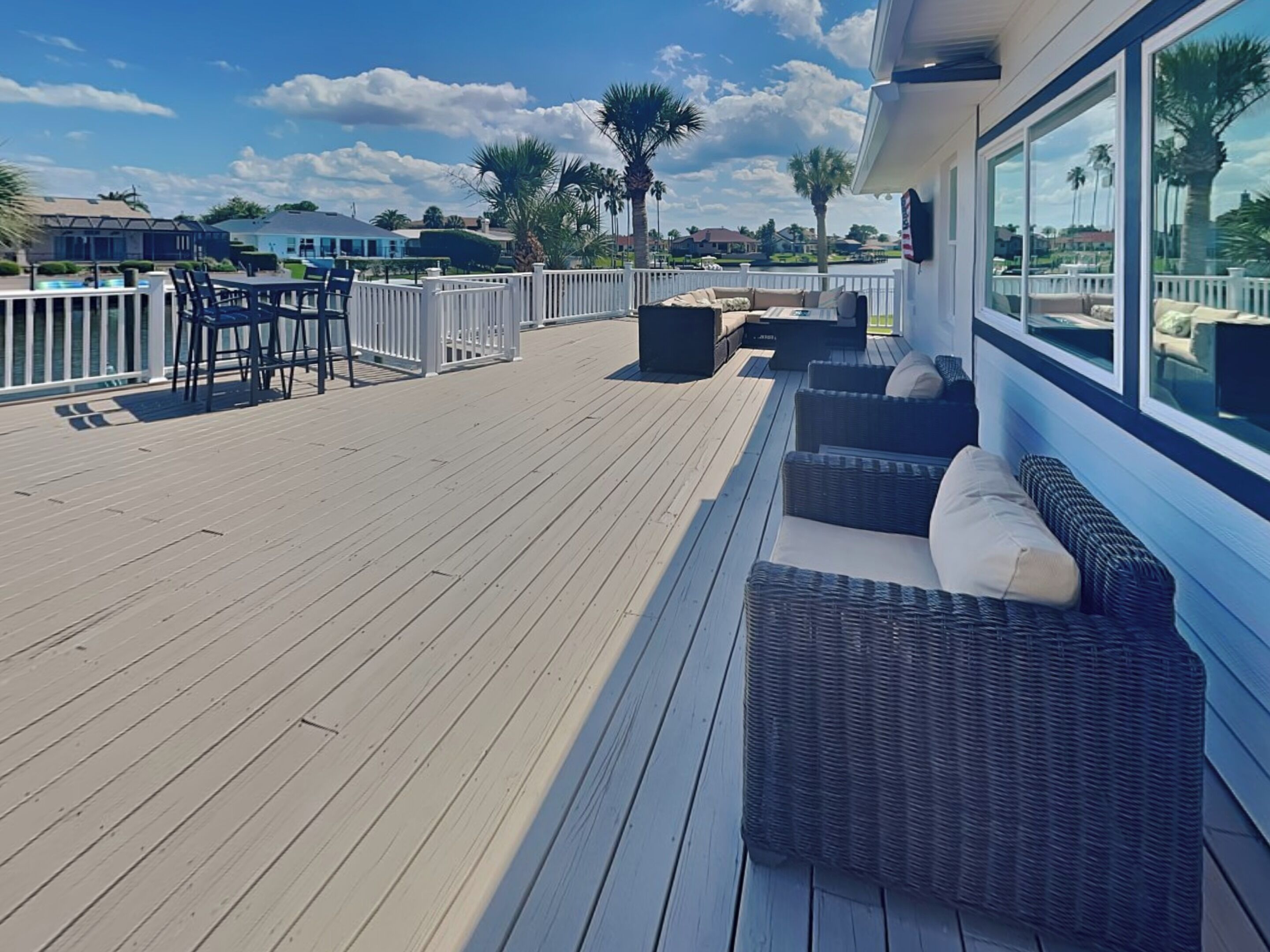A wooden deck with outdoor furniture, including a sofa and chairs, overlooking a residential area with palm trees and houses.